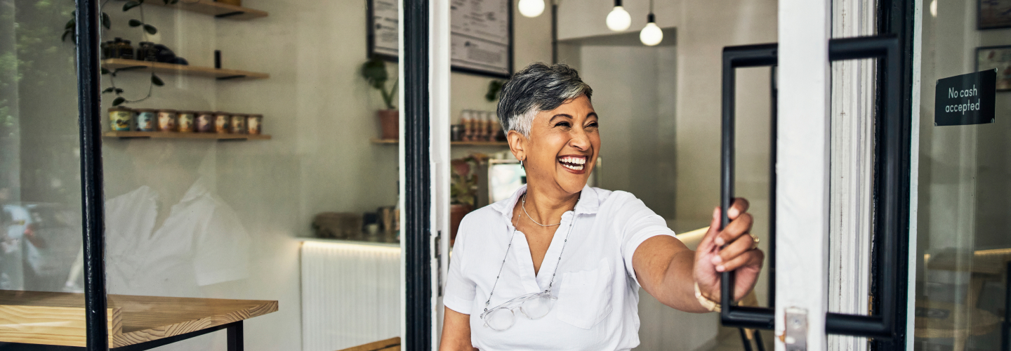 Smiling woman opening the door of a coffee shop, with shelves of coffee products and warm lighting visible in the background.