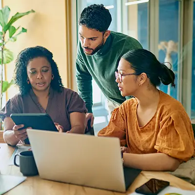 A diverse group of professionals, including a black woman, a man, and a woman, are working together in a modern office space. They are using a laptop and a tablet to collaborate on a project.