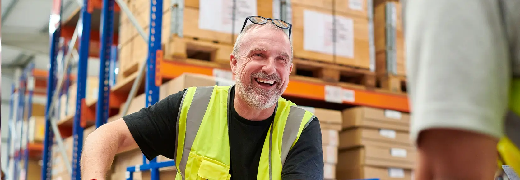 A middle-aged male warehouse worker with a gray beard and short hair is wearing a high-visibility yellow vest over a black t-shirt.